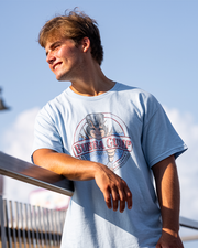 A young adult leans casually against a railing, wearing a light blue Bubba Gump Shrimp Company t-shirt, under a clear blue sky.