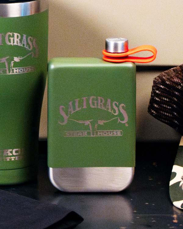 A green flask with a silver bottom and cap. The flask has the text "SALTGRASS STEAK HOUSE" printed on it. There is also an orange strap attached to the cap. The background includes other items, such as a green tumbler with similar branding and some dark-colored objects.