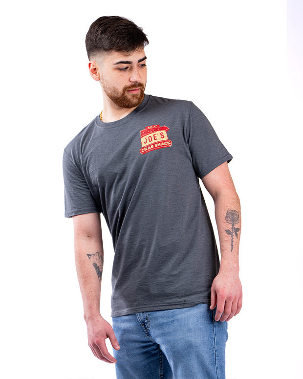 The image features a person wearing a grey t-shirt with a “Joe’s Crab Shack” logo on the left chest area, paired with blue jeans. They are standing against a plain white background.