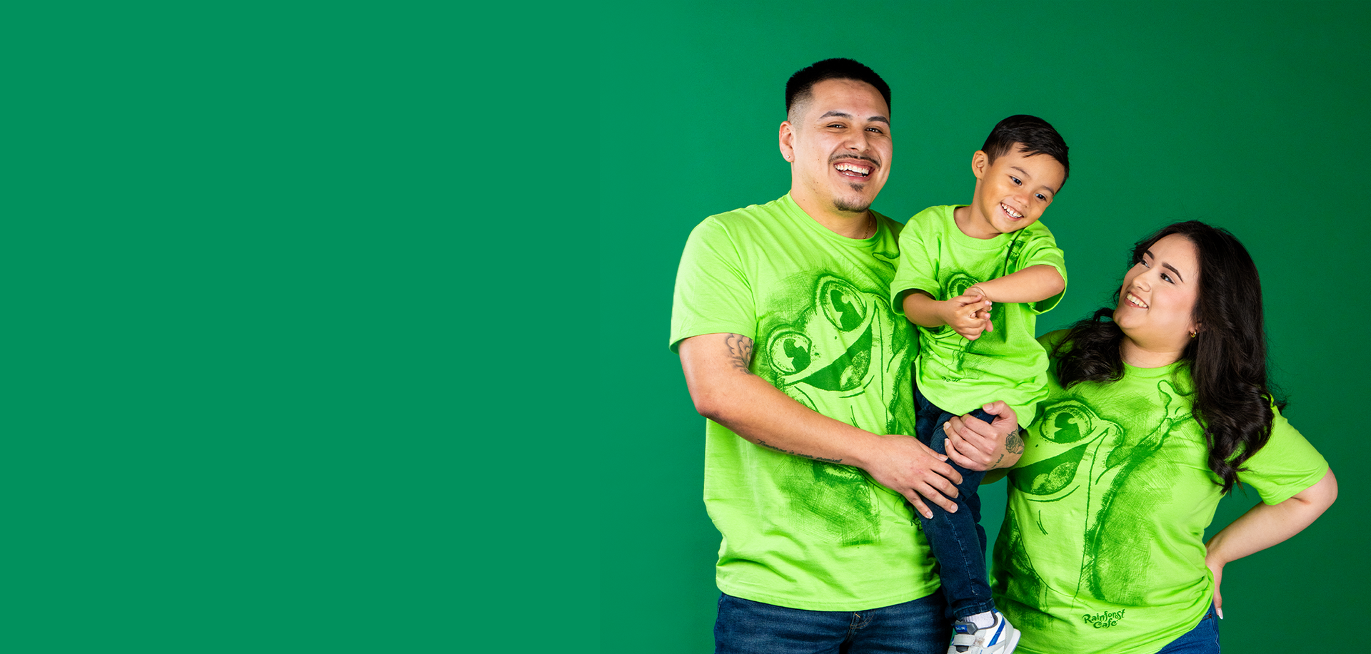 A family wearing bright green shirts stands together in front of a solid green background, showcasing playful smiles and unity.